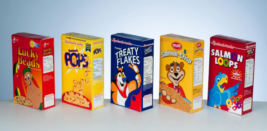 Five brightly coloured cereal boxes reinterpret familiar breakfast packaging with Indigenous-inspired imagery and playful, satirical graphics.