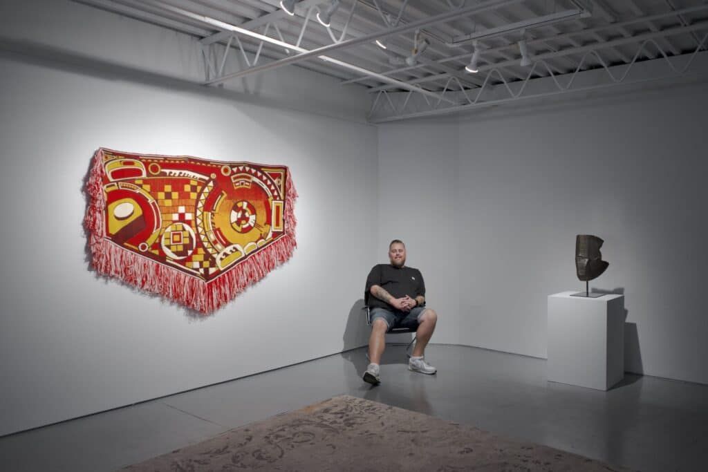 A person sits quietly in a gallery between a vibrant woven textile hanging on the wall and a sculptural work on a pedestal, surrounded by open space that invites reflection on the artworks.