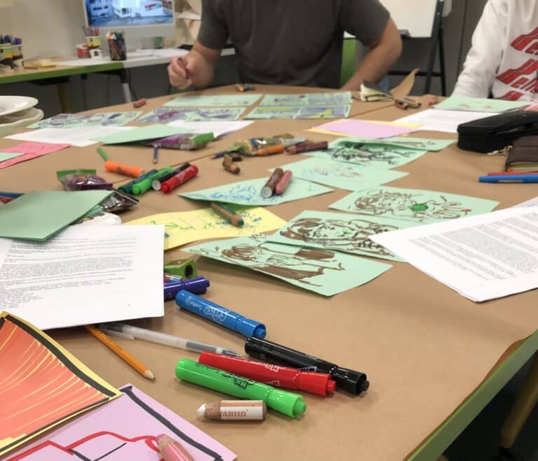 Markers, papers and illustrated drawings cover a large table as students work together on a collaborative classroom art activity.