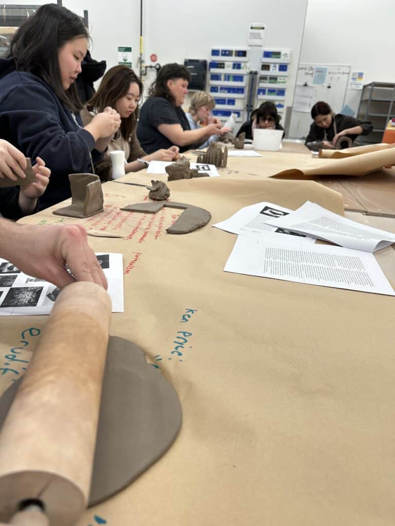Students shape clay at a large studio table during a hands-on ceramics workshop while reviewing printed course materials.