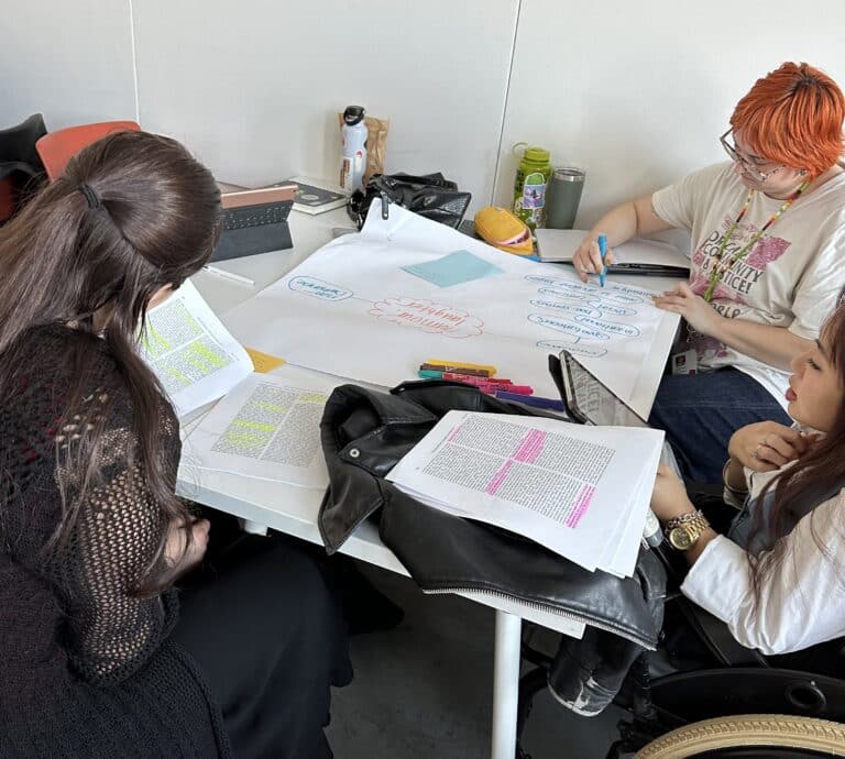 Students gather around a table highlighting readings and creating a collaborative concept map on a large sheet of paper.
