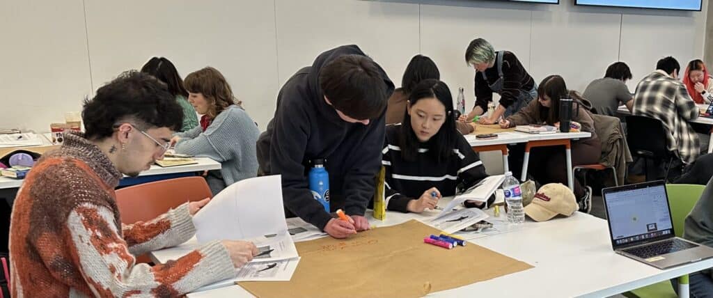 Students work in small groups during a seminar, reading course materials, writing notes and discussing ideas around classroom tables.