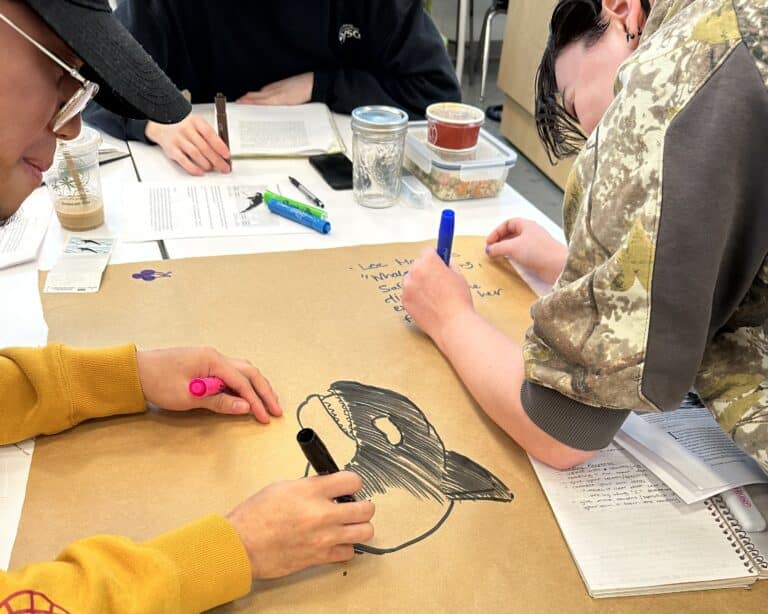 Students draw and write on a large sheet of paper while referencing course readings during a collaborative classroom activity.