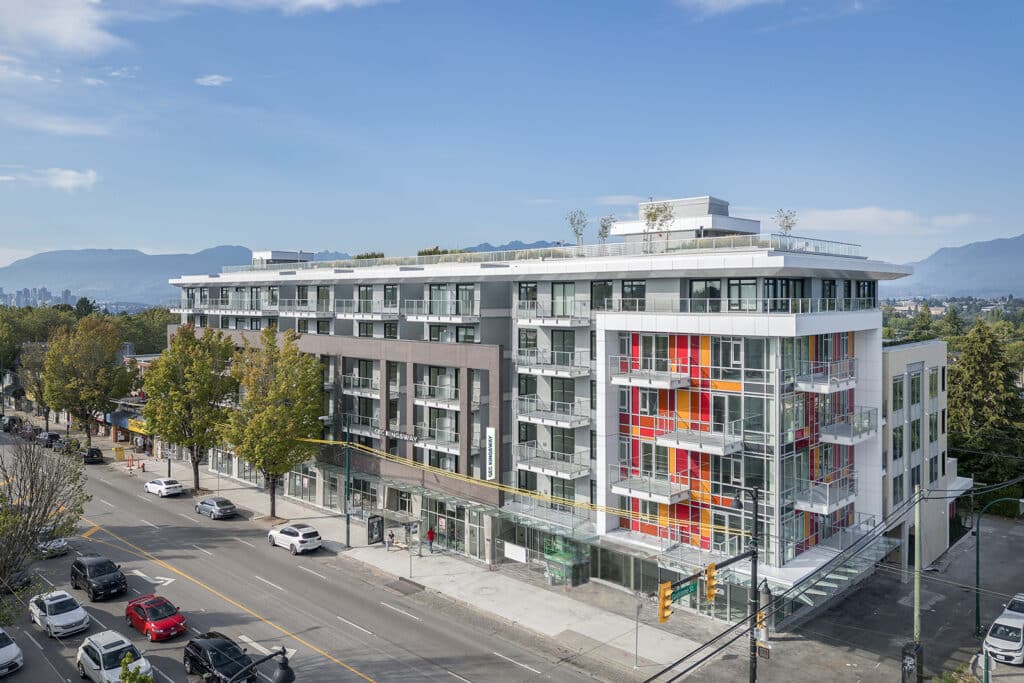 A modern six-storey mixed-use building sits on a street corner, featuring glass balconies, large windows and a colourful red, orange and yellow glass façade. Trees line the sidewalks, cars travel along the surrounding streets and mountains rise in the distance beneath a partly cloudy sky.
