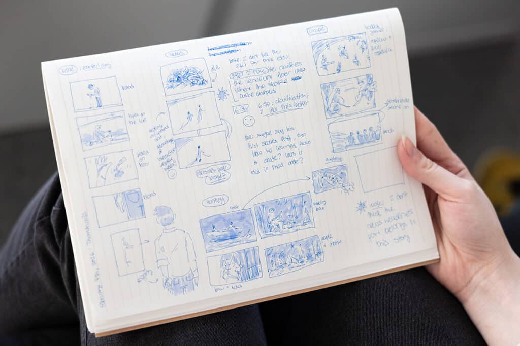 Person holding an open sketchbook filled with hand-drawn storyboard panels, notes and annotations in blue ink.