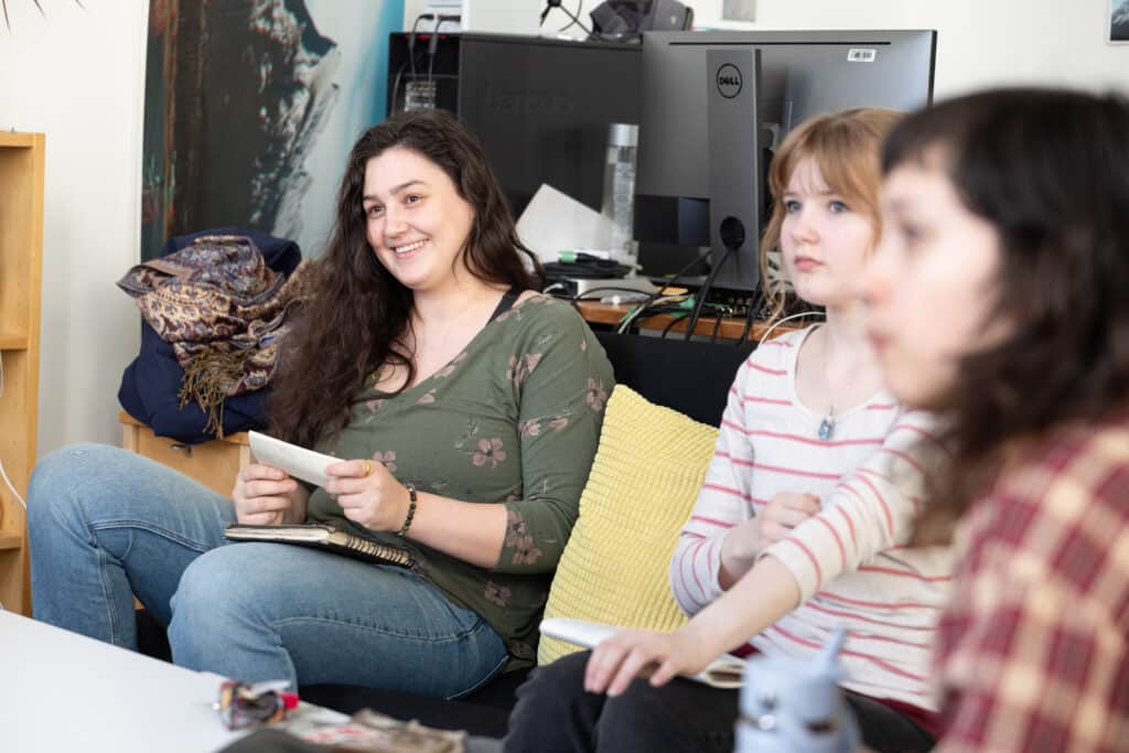 Small group seated in a studio classroom, listening and smiling during a discussion while holding notebooks.