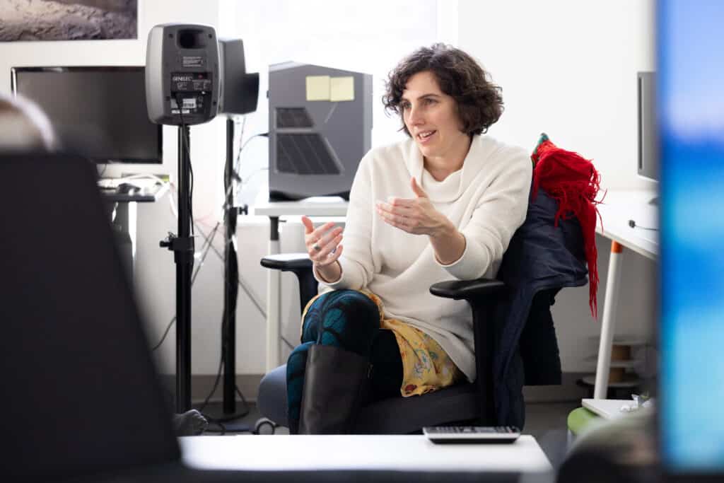 Person seated in an office chair gesturing with their hands while speaking during a group discussion in a media lab.