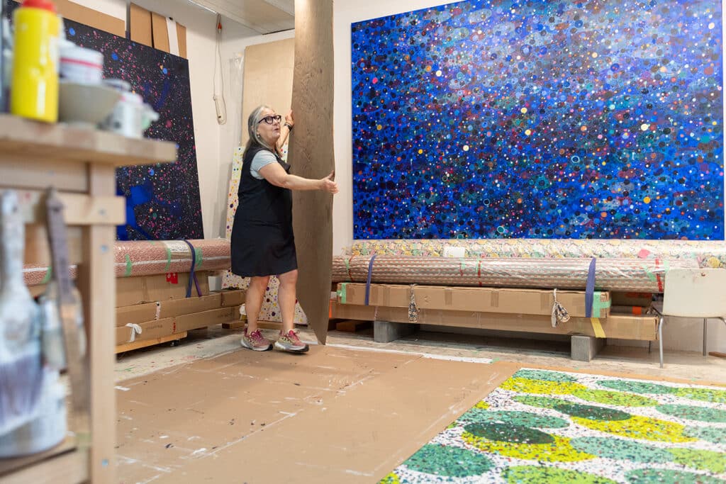 Person standing in an artist's studio holding a large cardboard panel beside colourful abstract paintings and wrapped canvases.