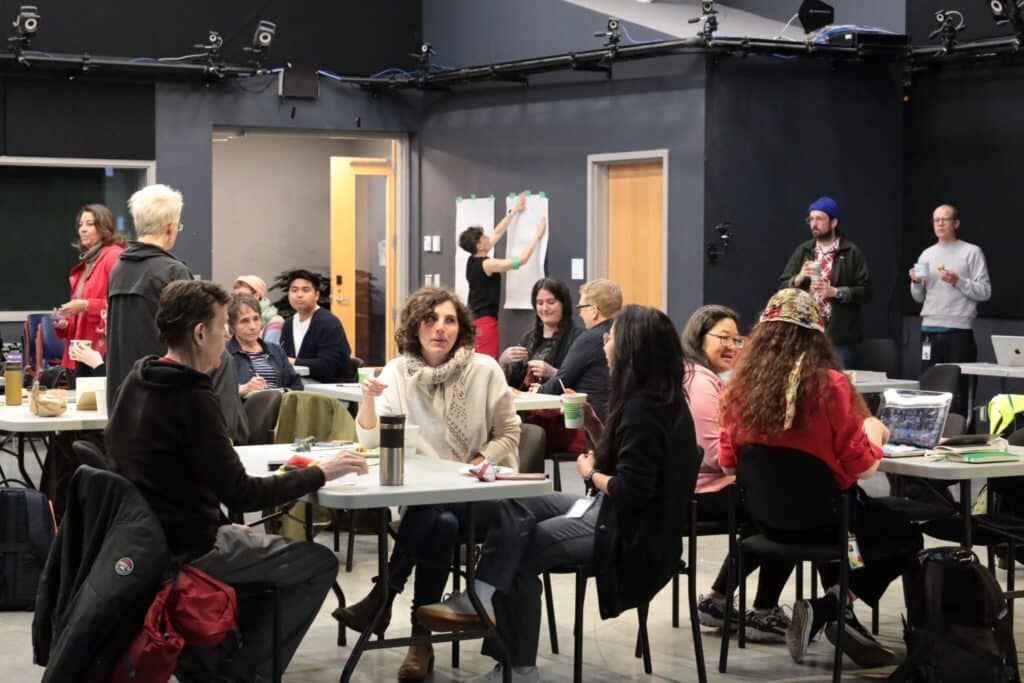 Participants gather around tables in conversation while others post notes on large sheets of paper during a collaborative symposium session.