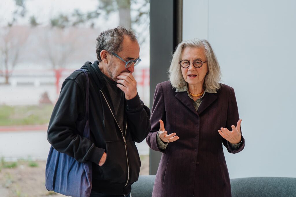 Two people converse beside a large window, one listening thoughtfully while the other speaks with open hands.