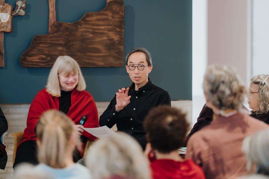 A speaker gestures mid-conversation during a panel discussion while audience members listen, creating a lively exchange of ideas beneath carved wooden artworks.