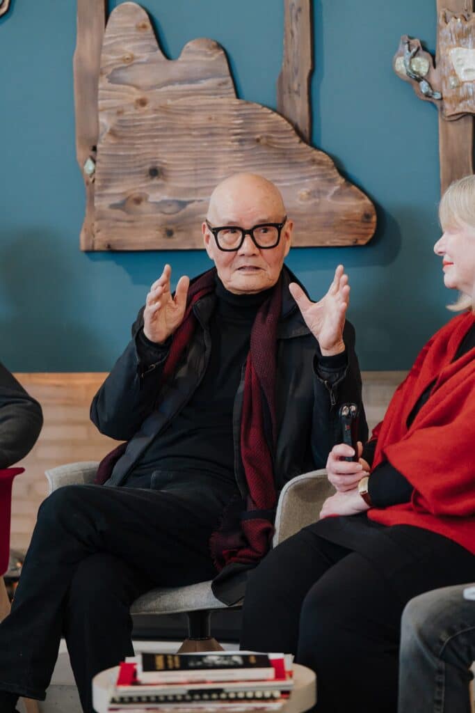 A speaker raises both hands while sharing a point during a panel discussion, framed by carved wooden artworks as fellow participants listen with focused attention.