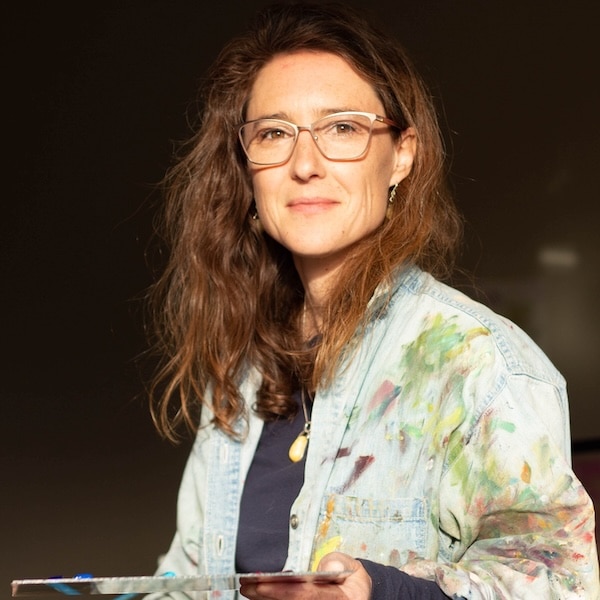 An artist stands in warm studio light wearing a paint-splattered jacket and holding a palette, with a calm expression that reflects a creative practice in progress.