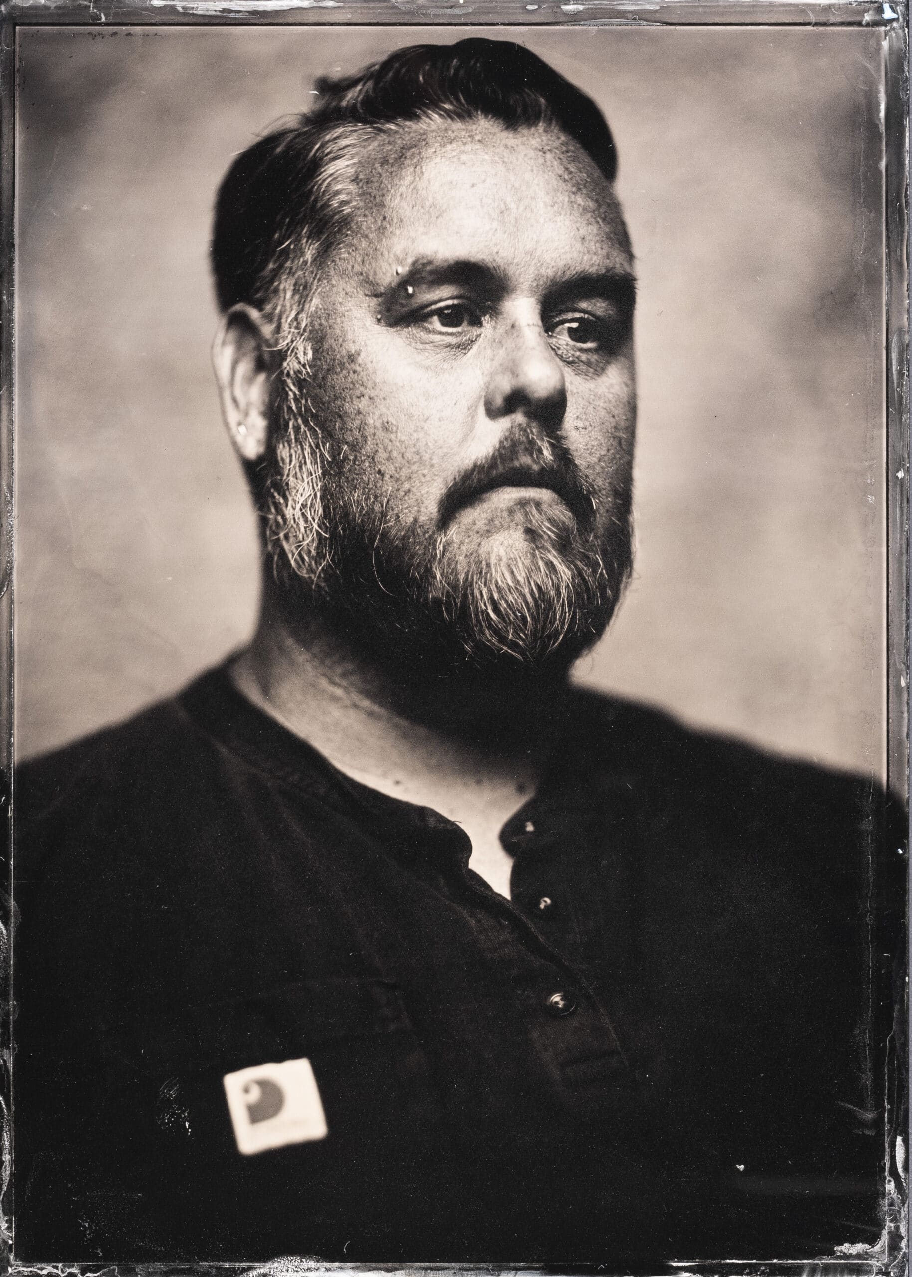 Sepia-toned portrait of a person with closely cropped hair and a full beard gazing thoughtfully beyond the camera, creating a calm and contemplative mood.