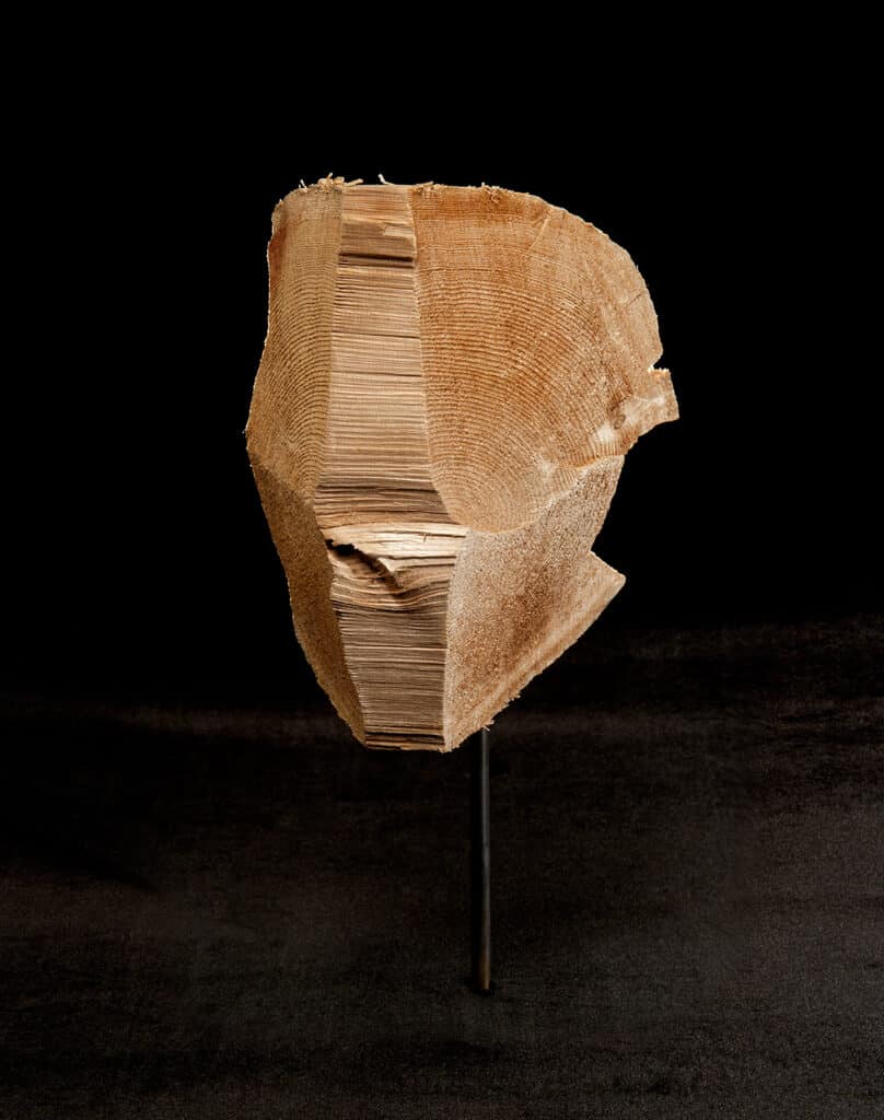 Carved wooden sculpture mounted on a slender metal stand, its rough-cut surface revealing layered grain against a dark background.