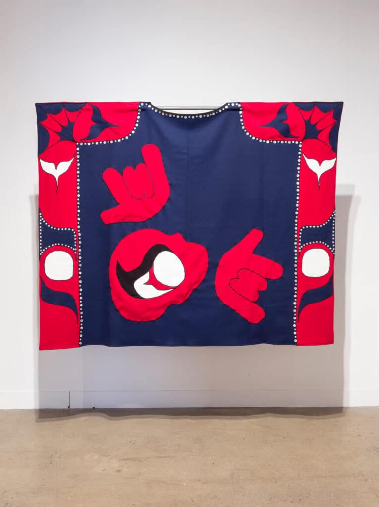 Bold red, navy and white textile hangs on a gallery wall, featuring stylized hands, eyes and floral motifs arranged in a balanced graphic composition.