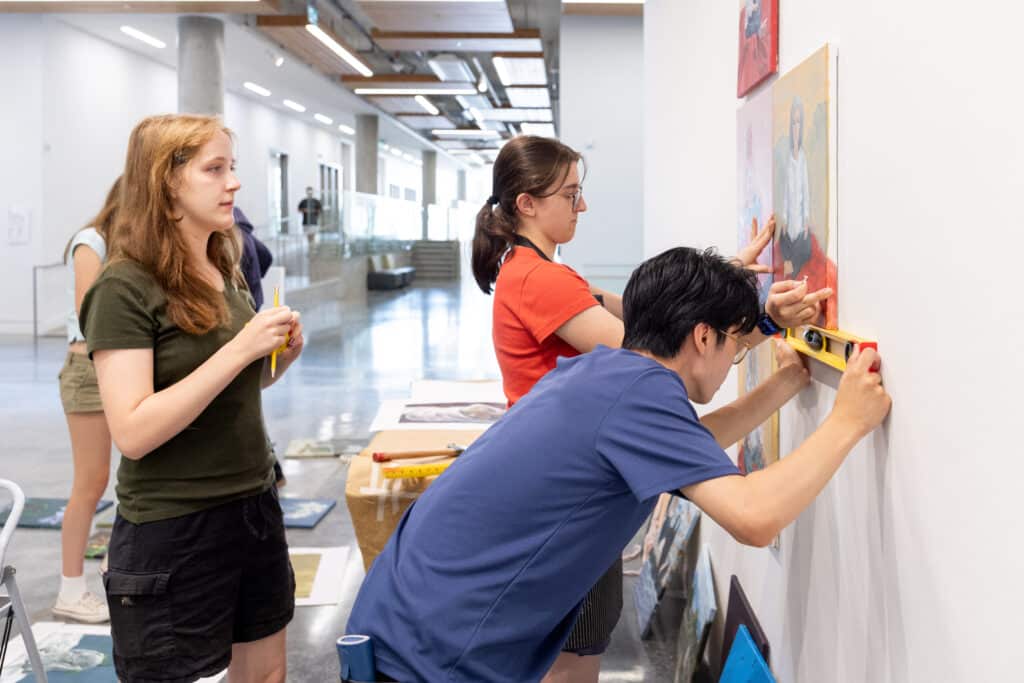 Three young artists use a level and measuring tools to position paintings on a gallery wall.
