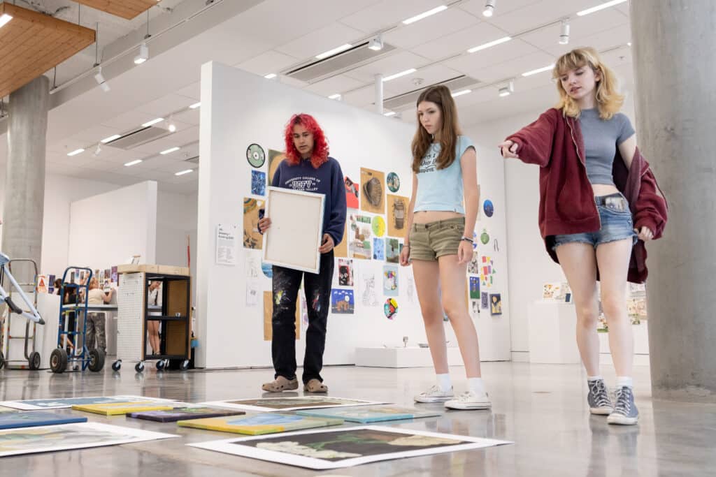 Three young artists review artwork arranged on the floor while planning an exhibition layout in a gallery.