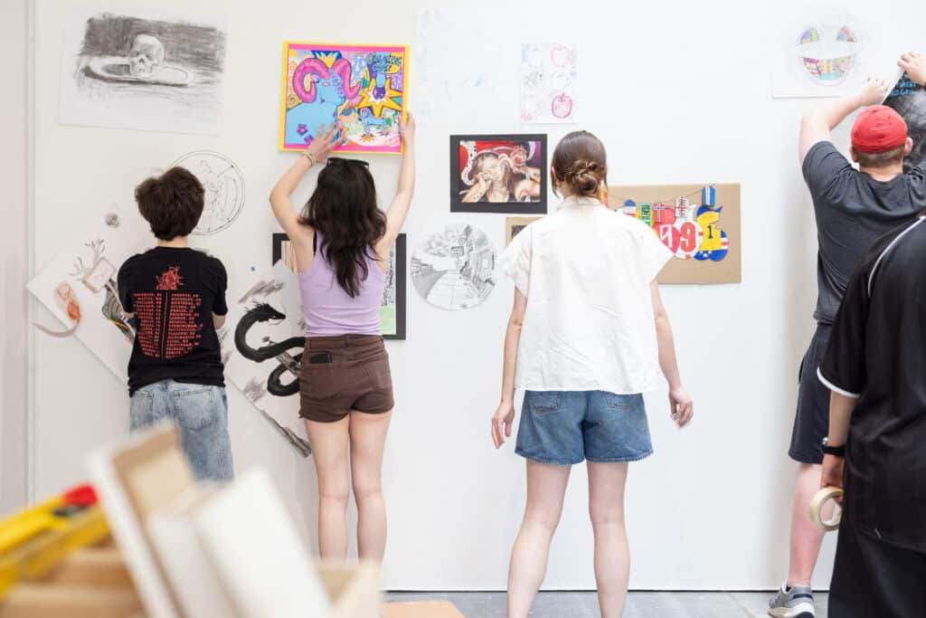 Students hang artwork on a gallery wall, arranging drawings, paintings and prints as they prepare an exhibition display.