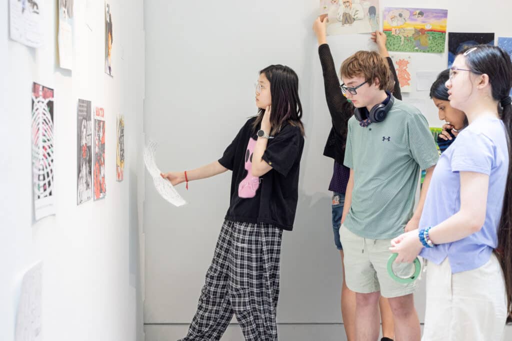 Students review, position and install artwork together on a gallery wall during exhibition setup.