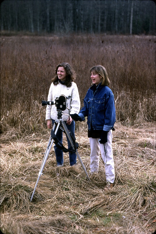 Two people stand beside a film camera on a tripod in a grassy field, with a forest in the background.