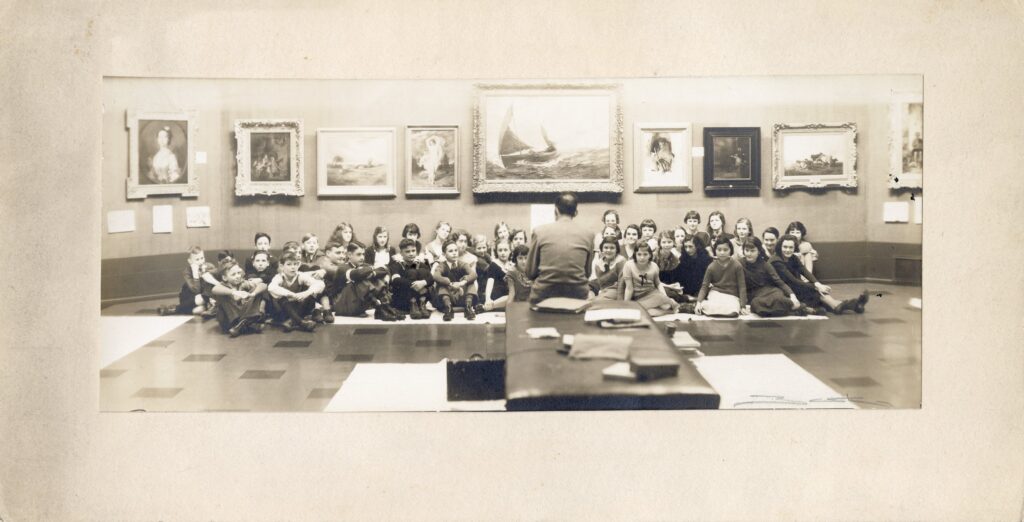 A group of children seated together in an art gallery as an instructor speaks to them, with framed artworks displayed on the wall behind them.