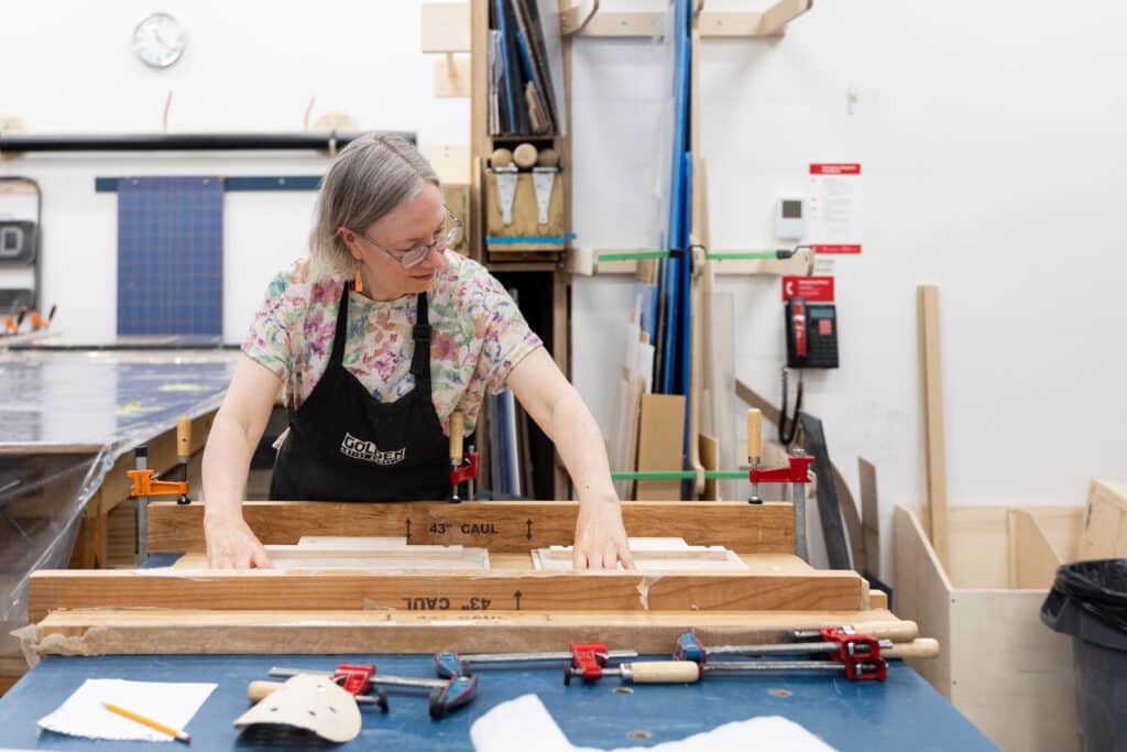 A person in a dark apron works with clamps, glue and wood on a large worktable in a brightly lit woodworking shop.