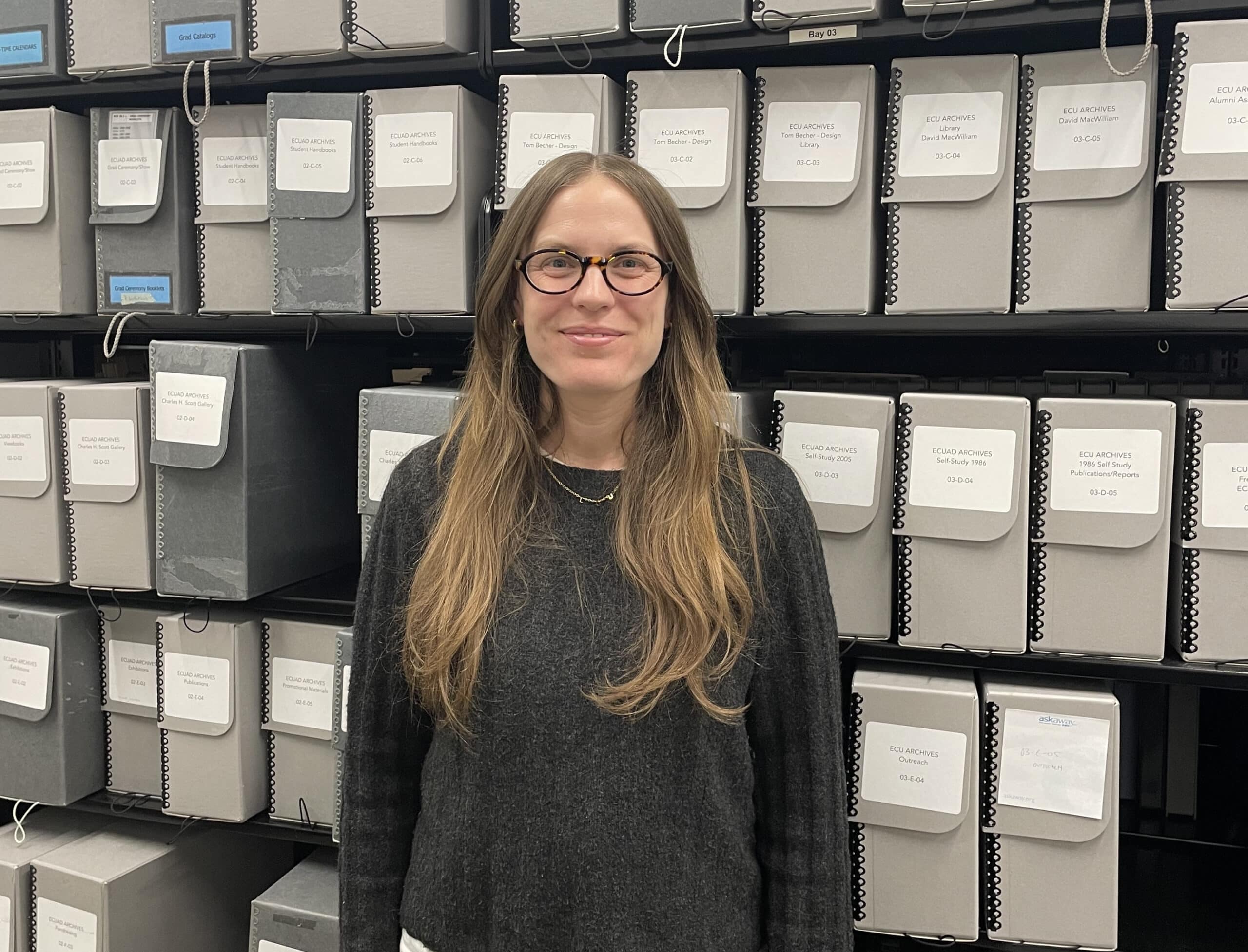A person with glasses stands in front of grey folders on a shelf