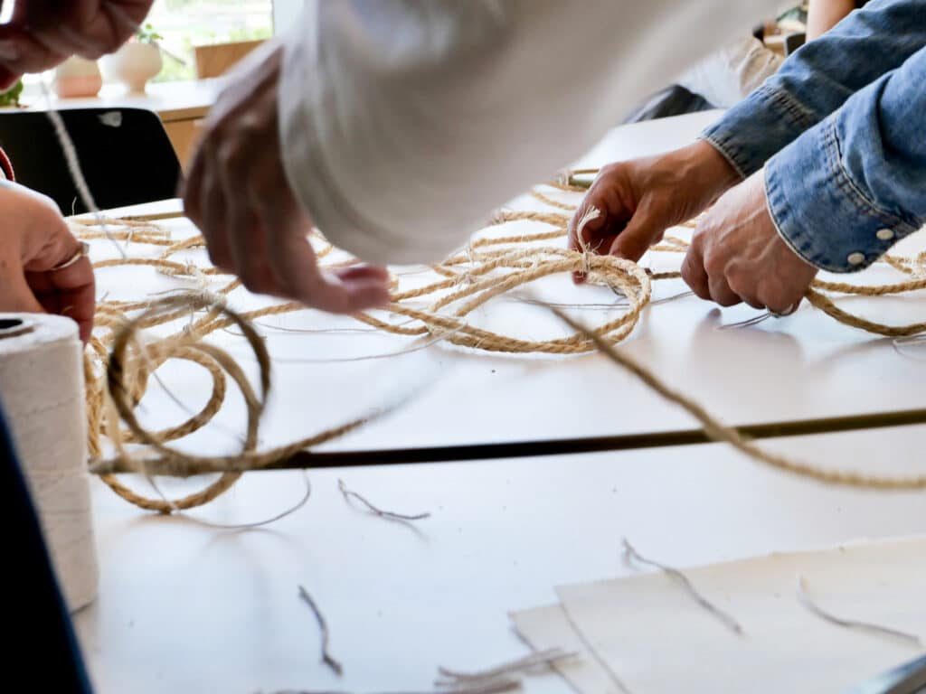 Several hands arrange and weave thick natural fibre rope across a table during a collaborative hands-on workshop.