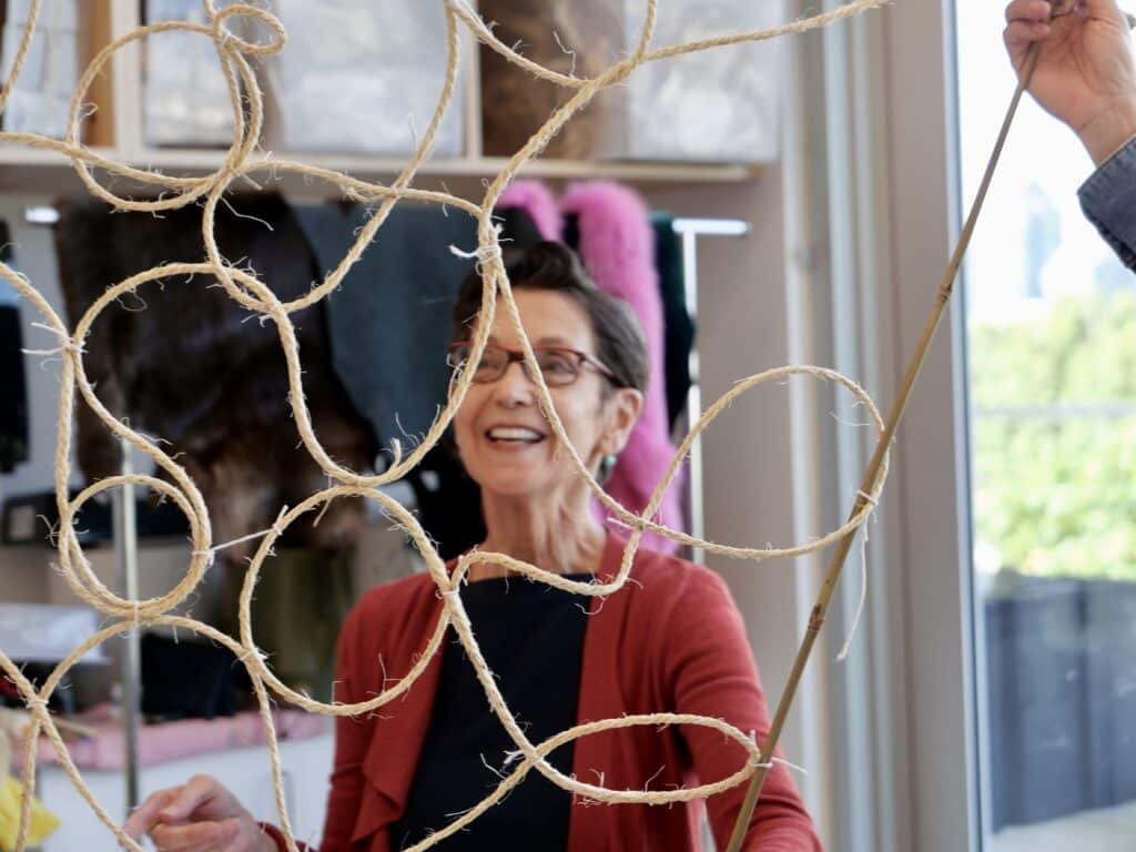 A workshop participant smiles while holding a reed structure intertwined with looping strands of natural fibre, with textile works visible in the studio behind them.