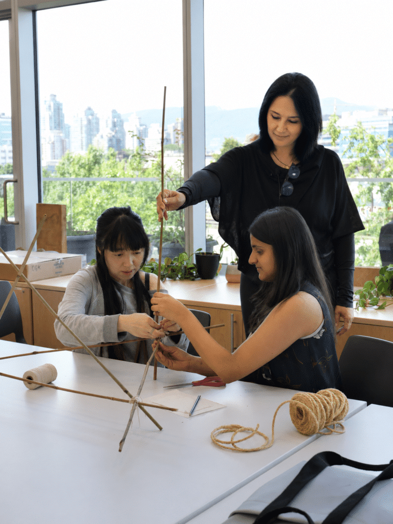 An instructor guides two participants as they assemble a sculptural framework from reeds and twine at a studio table overlooking the city.