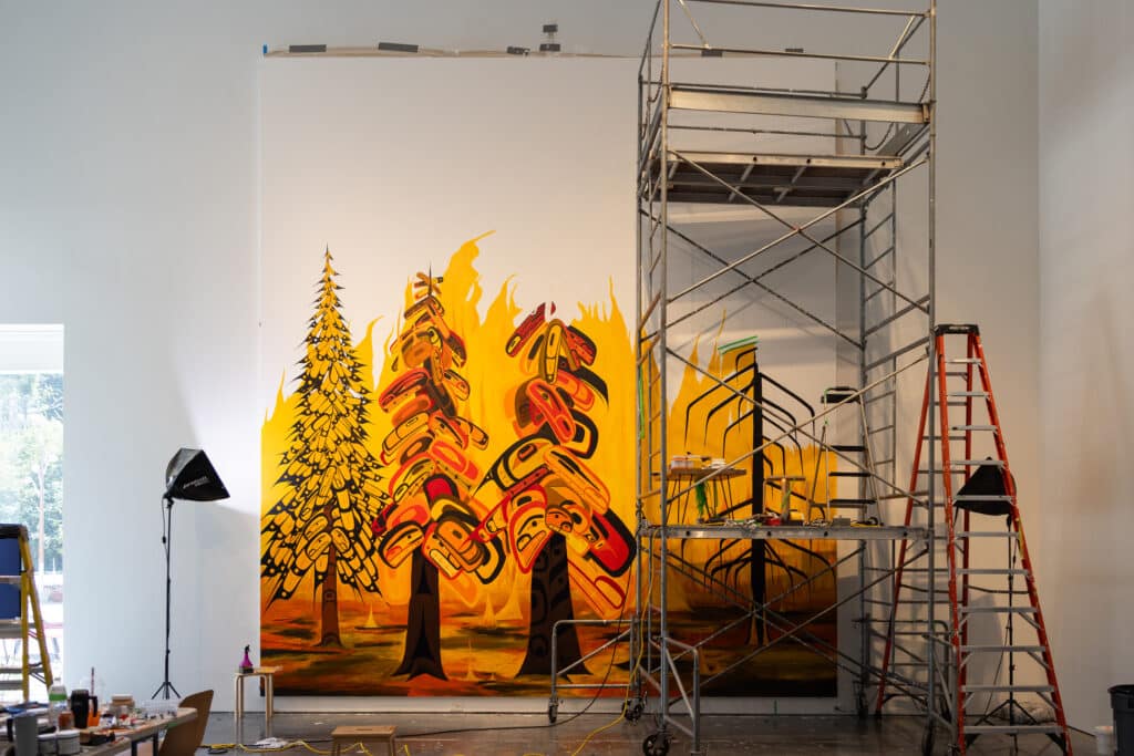 Large mural in progress inside a gallery, depicting stylized forest imagery in yellow, orange and black. Scaffolding, ladders and painting supplies surround the artwork.