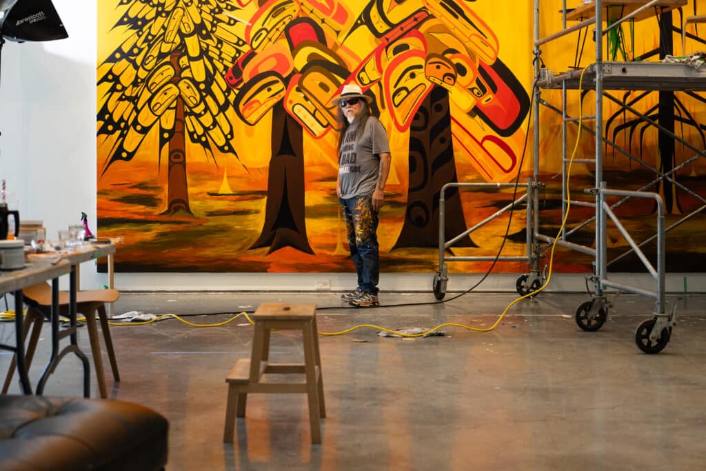 Wide view of an artist standing in front of a large mural in progress inside a studio, with scaffolding, extension cords and painting supplies visible.