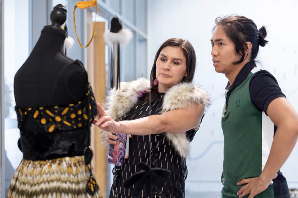 One person points out details on a garment displayed on a dress form while another looks on.