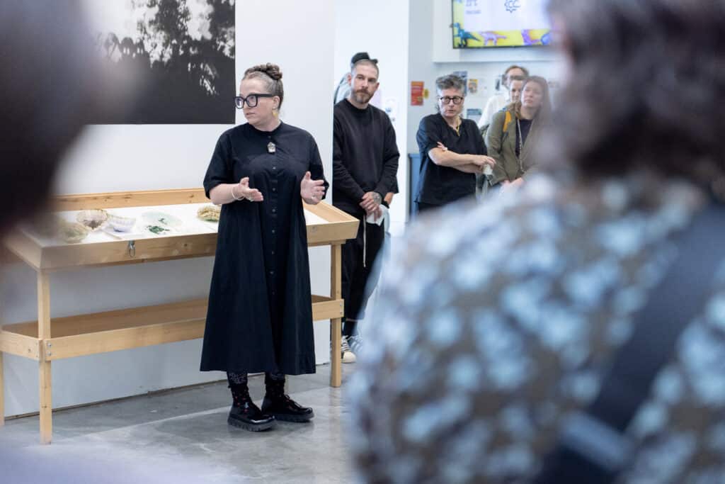 A speaker presents to an audience beside an exhibition display while visitors listen in the gallery.