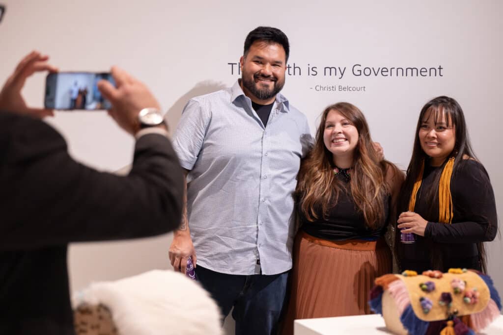 Three people pose for a photo in front of an exhibition wall while another person takes their picture.