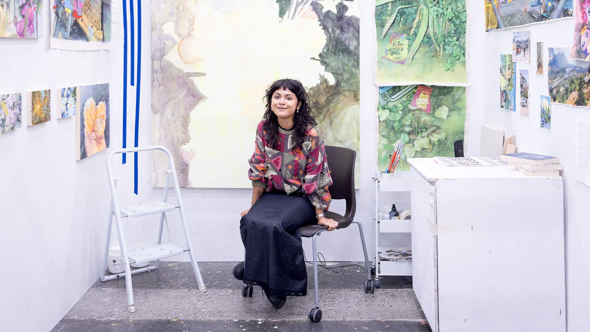 A student sits on a chair in their studio, surrounded by their paintings.