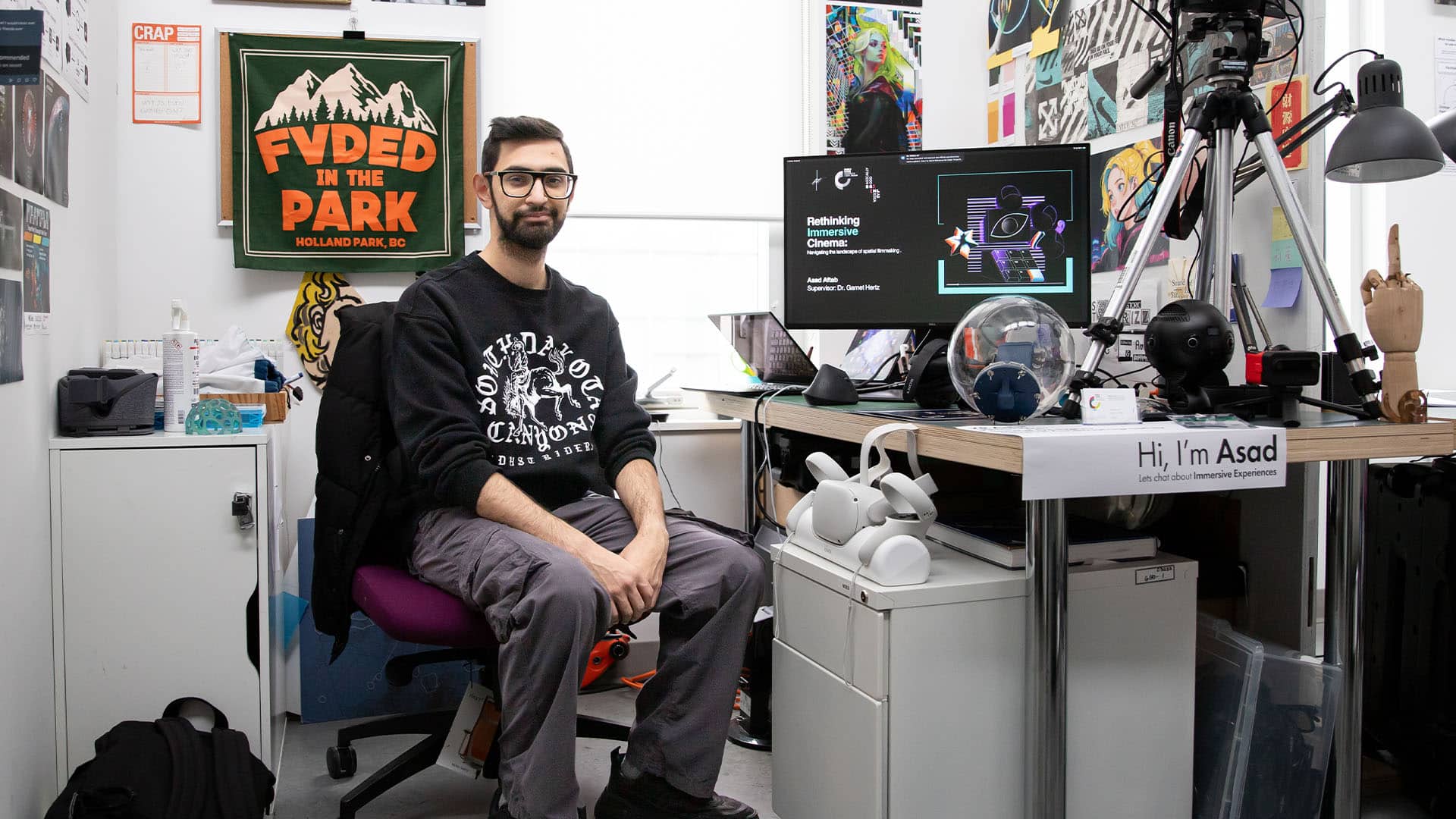 A grad student sits at his desk with a large monitor showing a project. The desk is full of interesting camera and tech equipment. He is facing the camera and smiling.