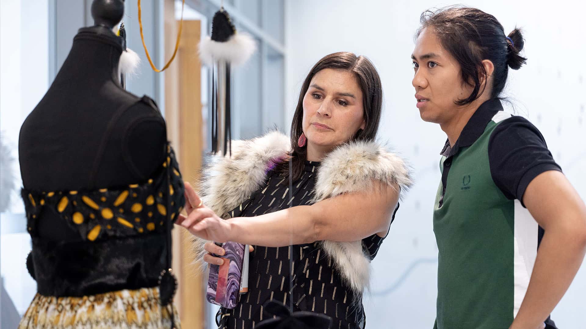 A student stands with another person in front of a dressmaker's mannequin, pointing out a detail on the garments on display.
