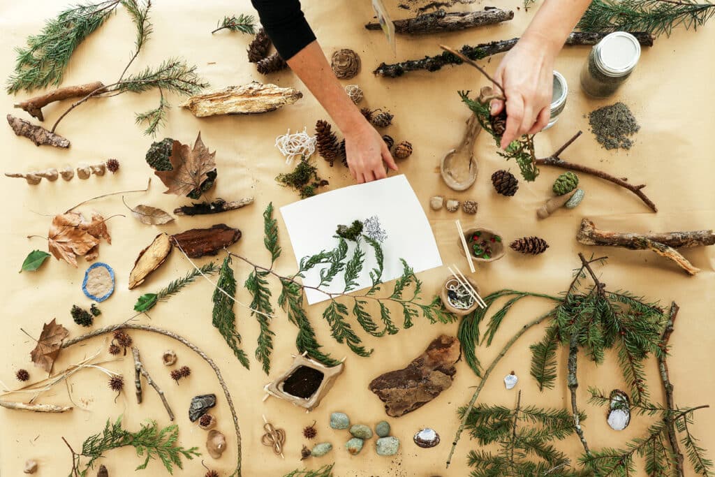 Hands arrange evergreen branches, pinecones and other natural materials across a table alongside small handmade objects and a botanical drawing.