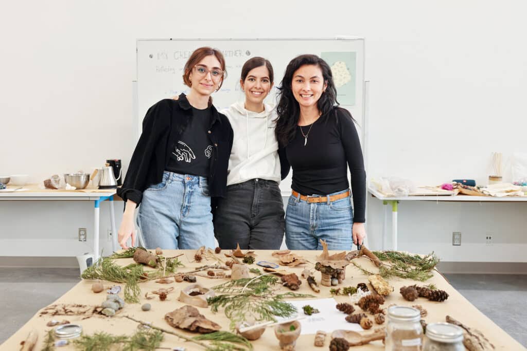 Three people stand together behind a worktable covered with evergreen branches, pinecones, stones and handmade objects in a classroom.