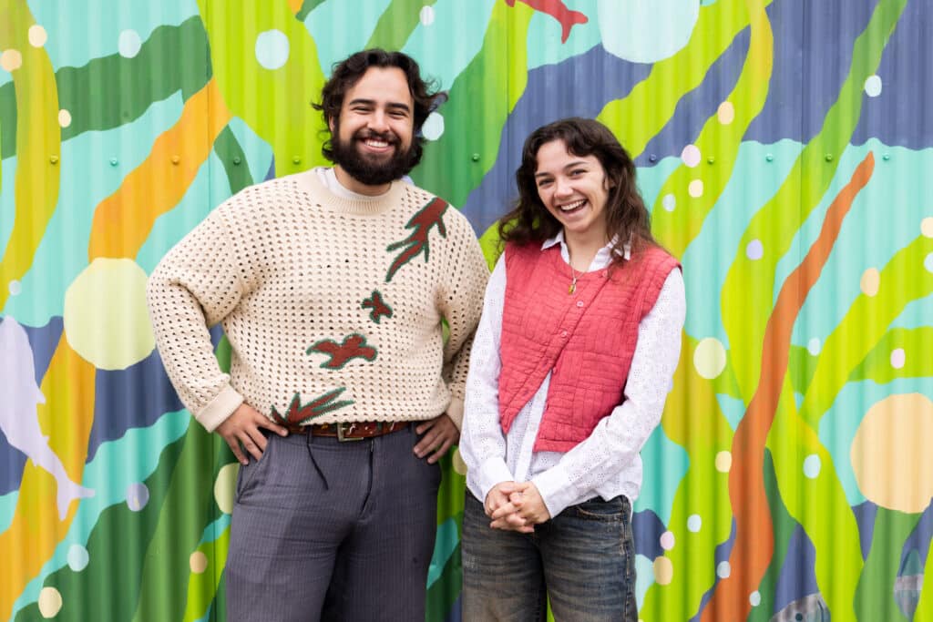 Two people smile in front of a vibrant mural featuring green eelgrass, orange strands and fish against a blue background.