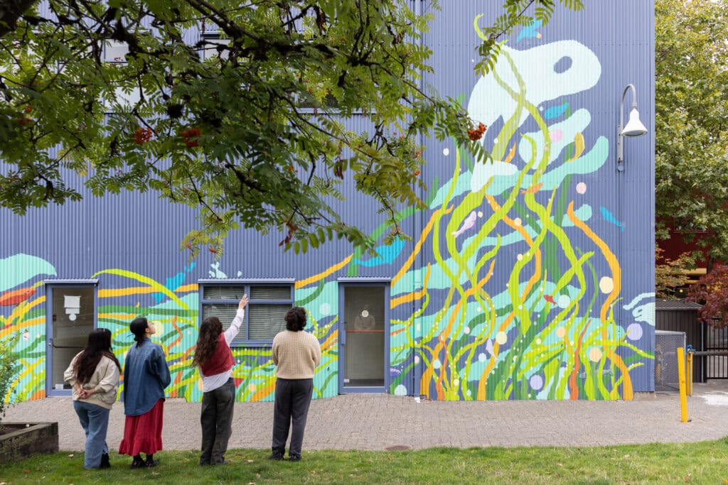 Four artists stand beneath a tree, looking up at a colourful aquatic mural. One points toward the artwork.