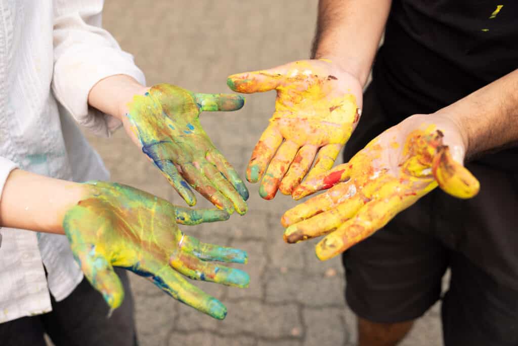 Four outstretched hands covered in vibrant yellow, green, blue and orange paint.