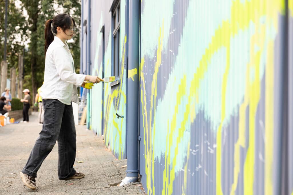 An artist wearing a white shirt and glasses paints a yellow line on a turquoise and blue mural.