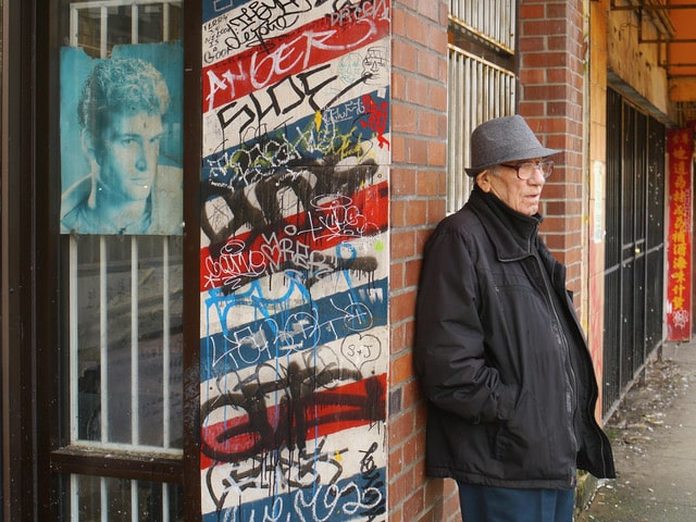 A person wearing a grey hat and dark coat stands beside a brick wall covered in layers of colourful graffiti.