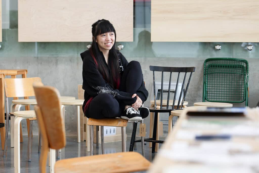 A person with long dark hair and pink highlights sits among an assortment of wooden chairs and stools in a bright studio.