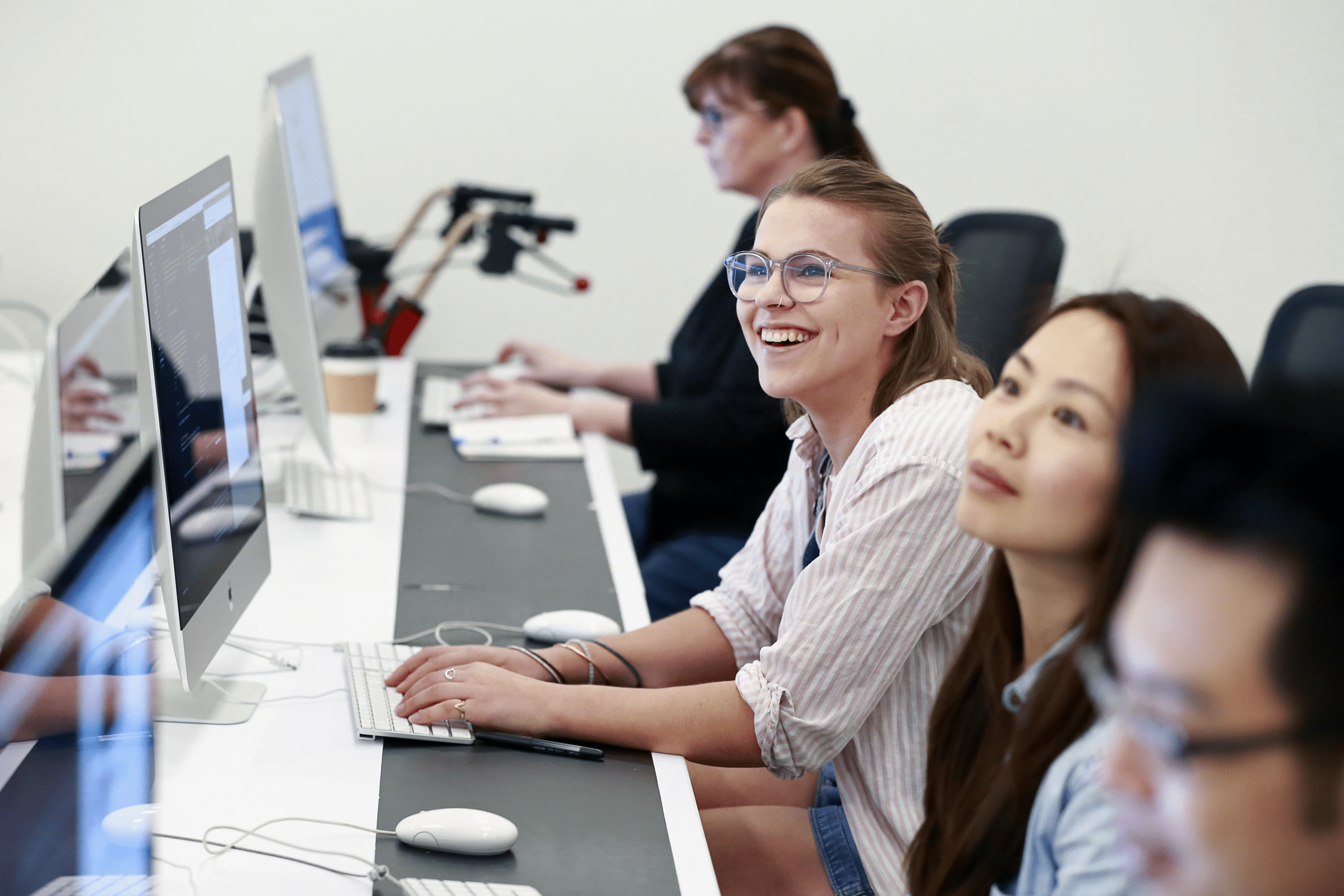 Students work at desktop computers in a bright computer lab, with one smiling student looking toward someone off-camera.