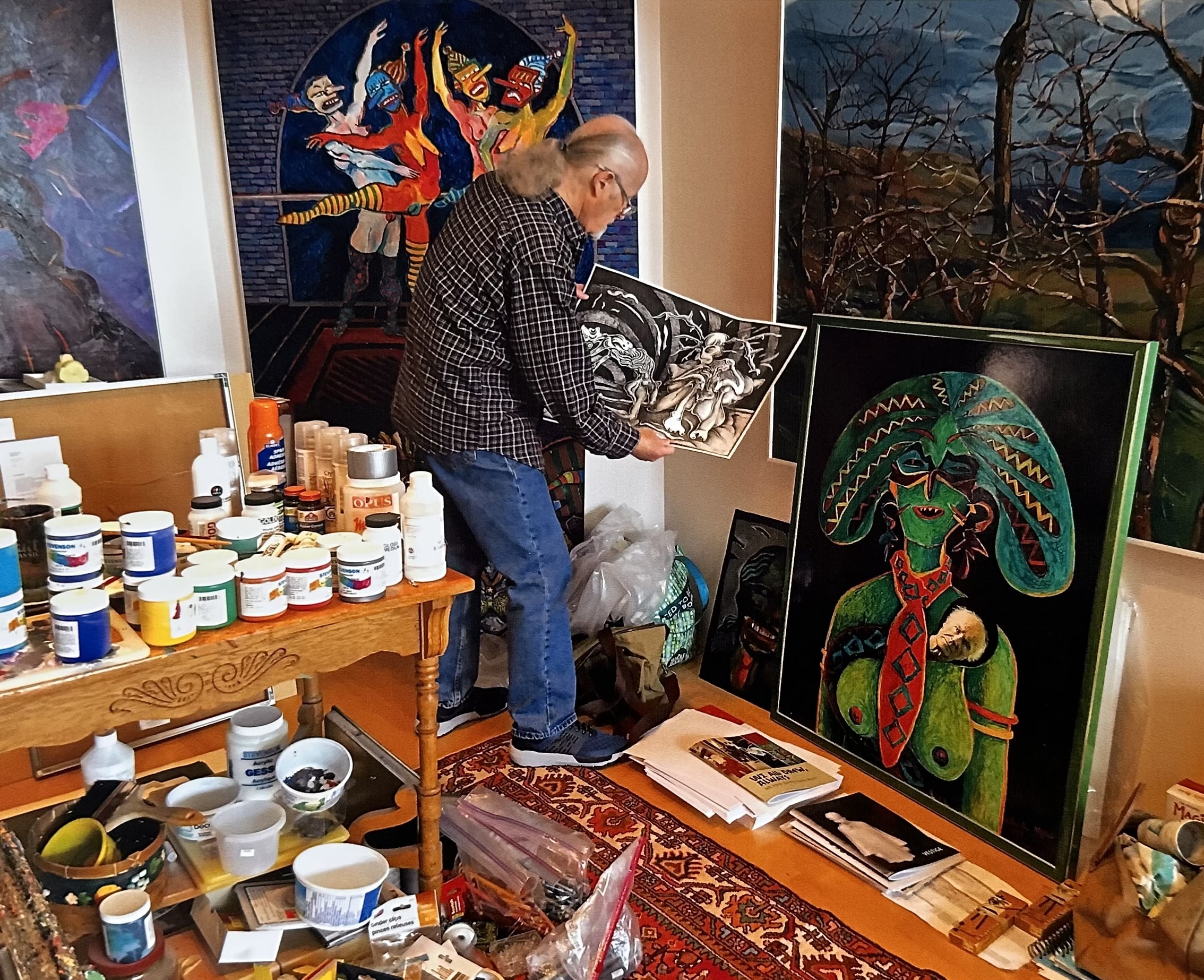 An artist stands in a crowded studio holding an artwork, surrounded by paintings, art supplies and containers of paint.