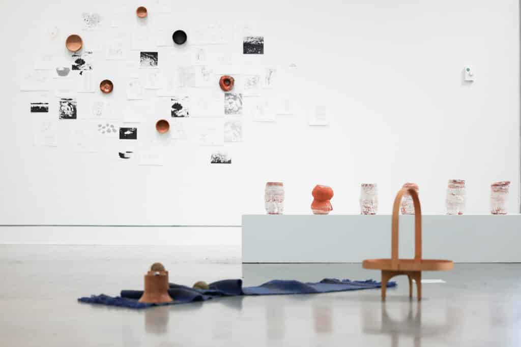 Gallery installation with ceramic vessels, drawings and printed works displayed across a white wall, plinths and the gallery floor.